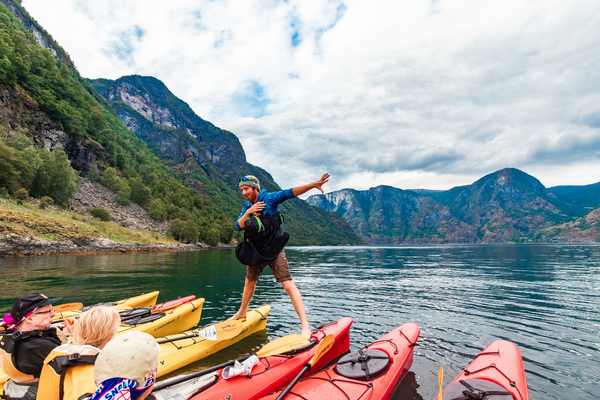 En mann balanserer lekent på tvers av en gul og en rød kajakk på en rolig fjord, til underholdning for andre padlere med bratte fjell i bakgrunnen.