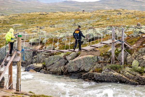 To turgåere med ryggsekk krysser en smal hengebru av tre over en brusende elv i et steinete fjellandskap.