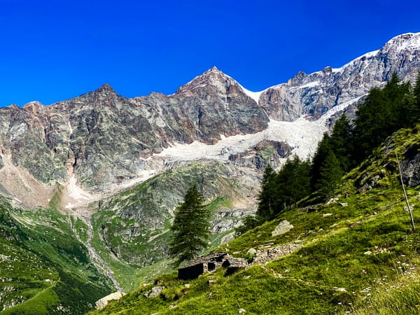 En liten, rustikk steinhytte ligger på en grønn gressbakke nedenfor ruvende, steinete fjelltopper og en hvit isbre under en klar, blå himmel.