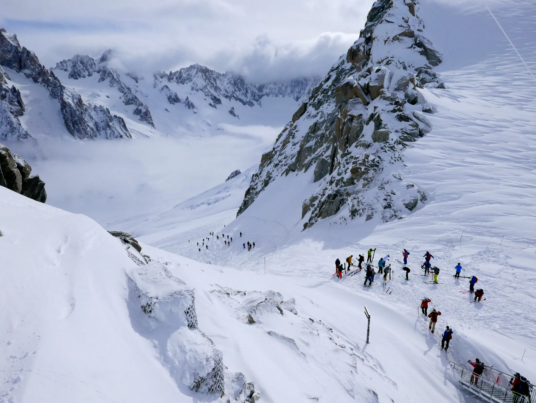 Tallrike skiløpere og fjellklatrere samler seg på et snødekt fjellpass flankert av bratte klipper, med et hav av skyer som fyller dalen nedenfor.