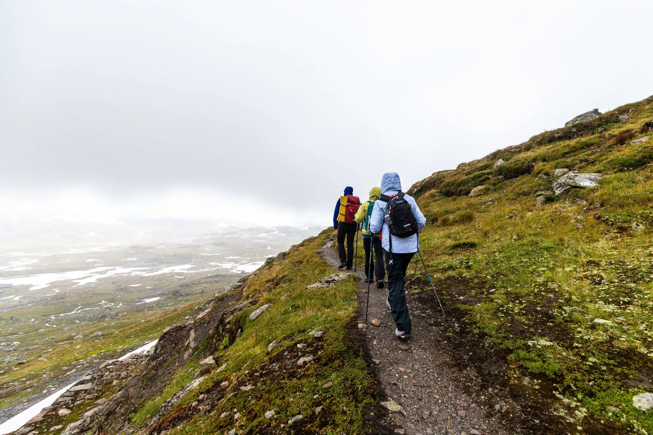 Tre turgåere med sekker og turstaver går langs en smal fjellsti på en tåkete dag, med utsikt over en stor, åpen dal.