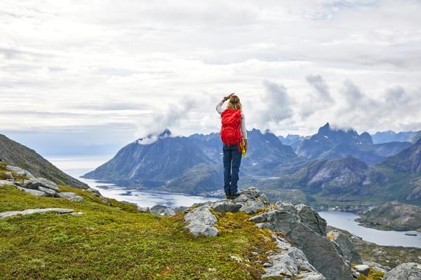 En turgåer med rød sekk står på en fjelltopp og ser ut over et fjordlandskap i Norge.