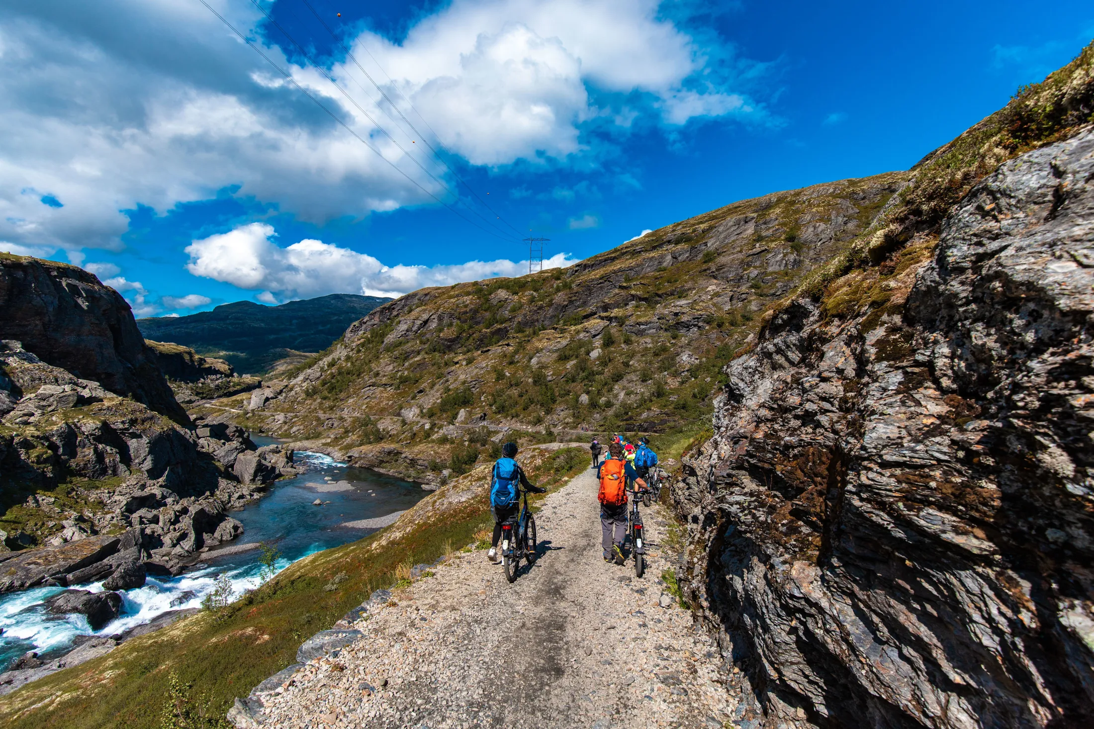 En gruppe syklister med fargerike sekker ferdes langs en grusvei ved siden av en brusende fjellelv under en klar, blå himmel.