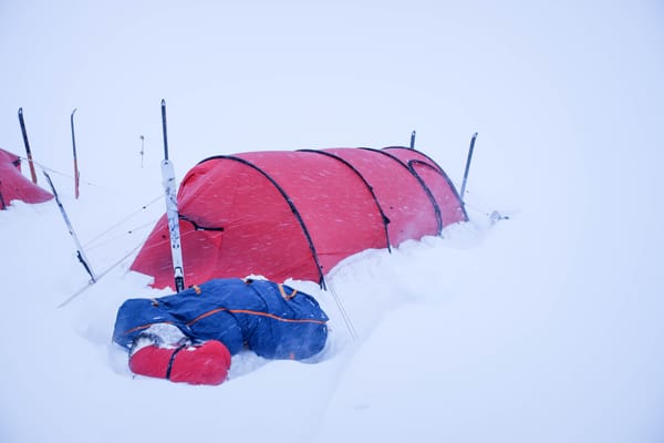 Et rødt tunneltelt slått opp i snøen under en snøstorm, sikret med barduner og loddrette ski, med en blå og rød pulkpose liggende i en snøgrøft i forgrunnen.