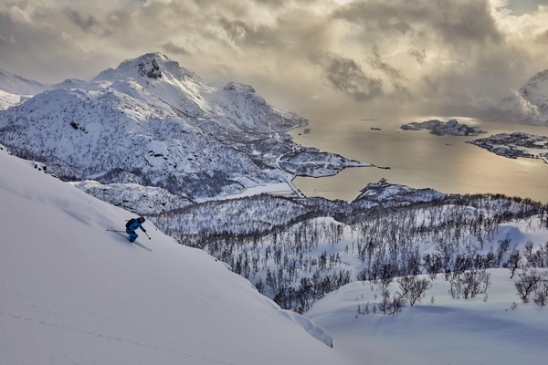 En skiløper renner ned en snødekt skråning med utsikt over en dramatisk fjord og fjell under en skyet himmel.