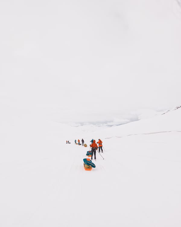 Skiløpere i oransje jakker trekker ekspedisjonspulker på rekke over et vaidt, hvitt arktisk landskap under en blek himmel.