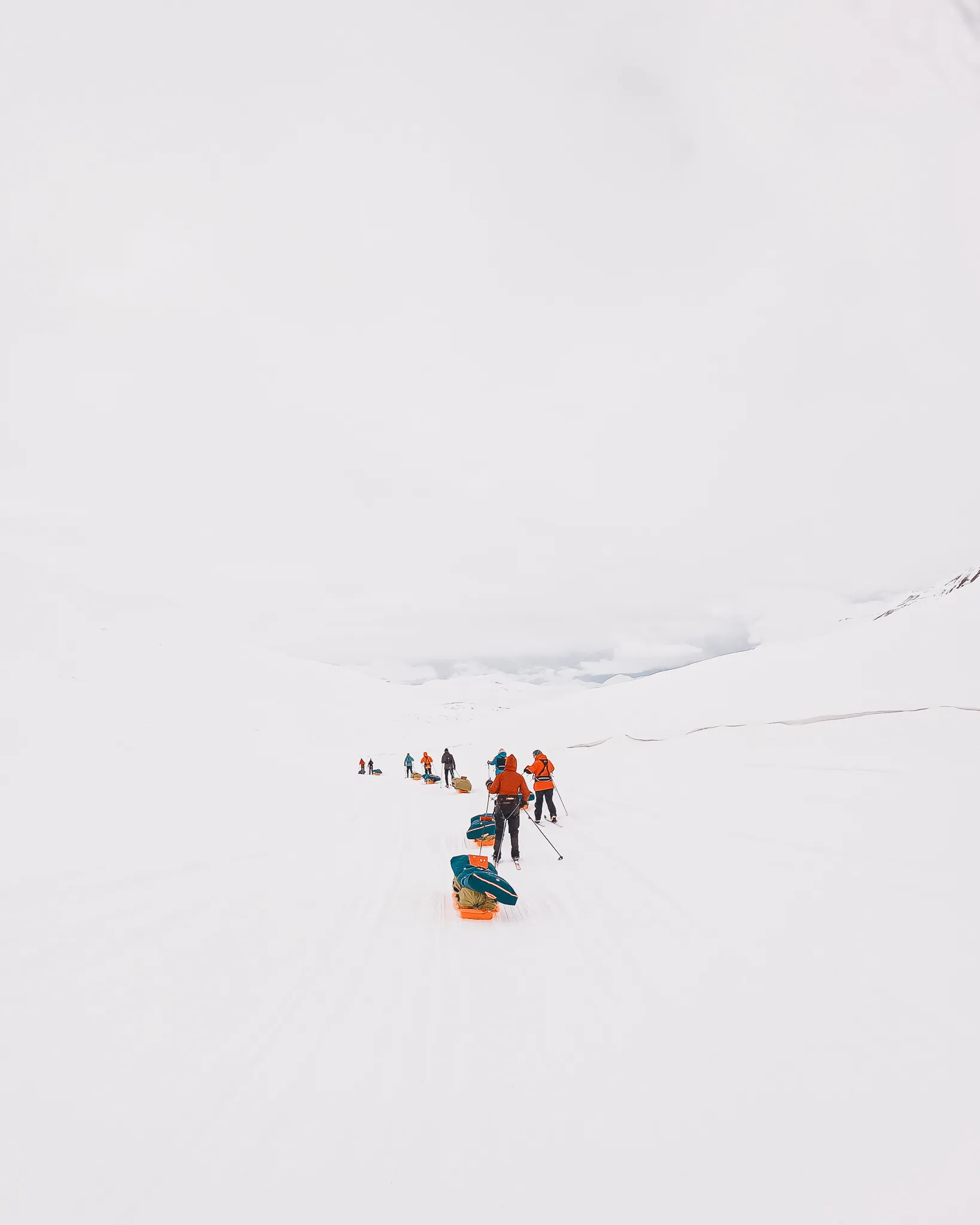 Skiløpere i oransje jakker trekker ekspedisjonspulker på rekke over et vaidt, hvitt arktisk landskap under en blek himmel.