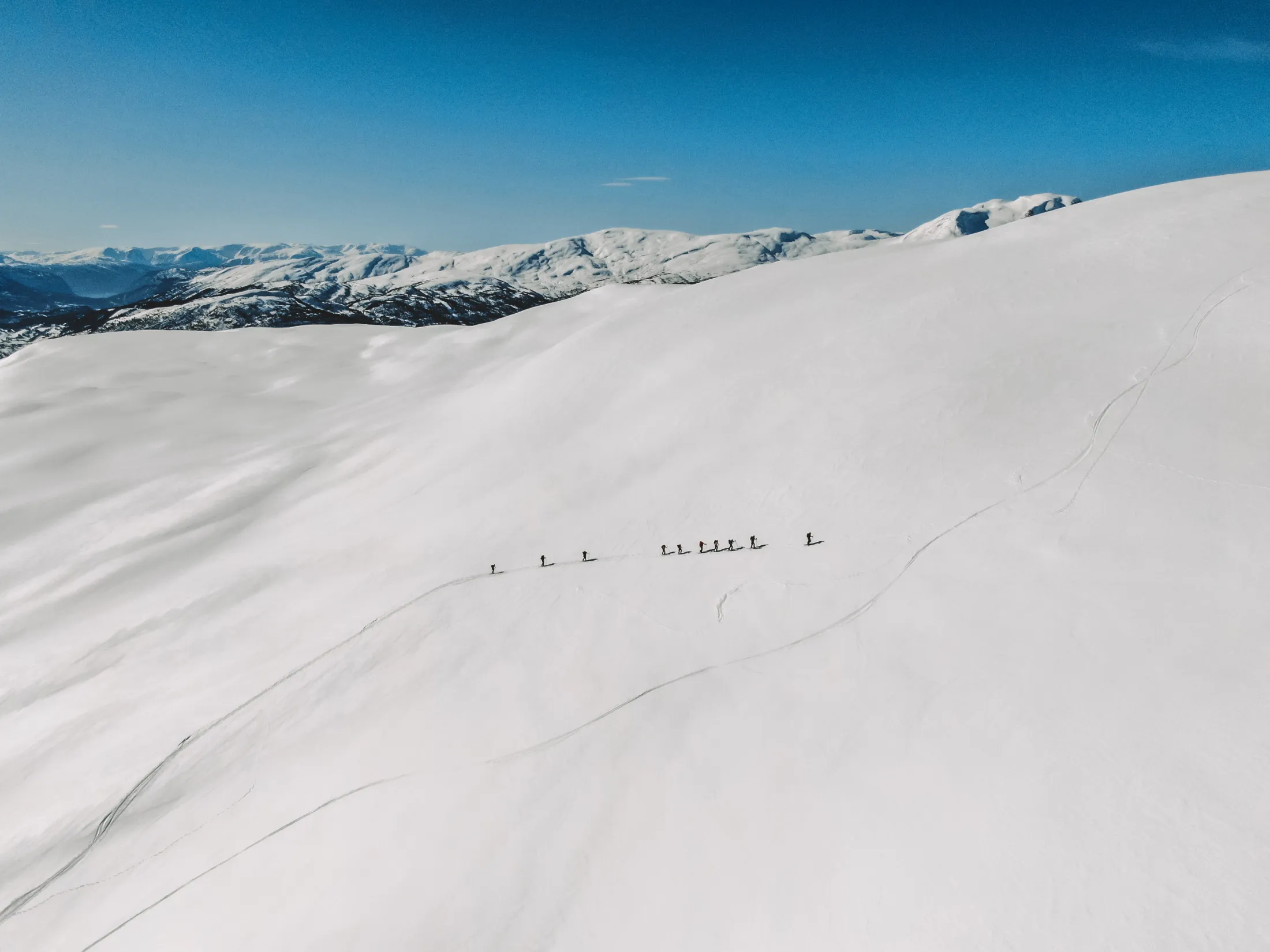 Et oversiktsbilde som viser en gruppe skiløpere som bittesmå figurer på en enorm, hvit snødekt fjellside under en klar, blå himmel.