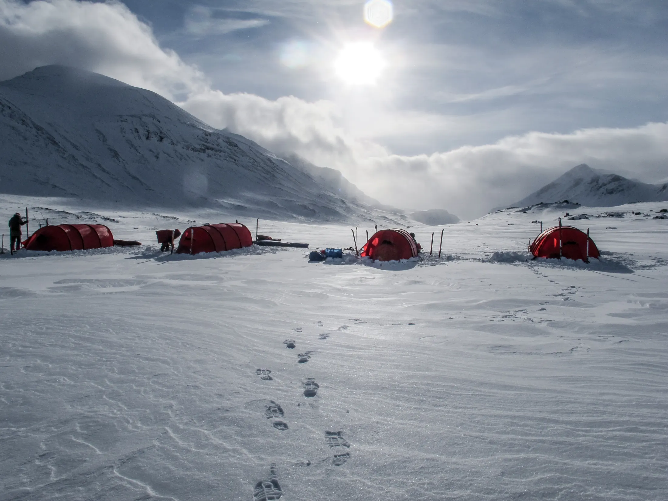 Røde telt satt opp i et snødekt fjelllandskap under en lys himmel med fotspor i forgrunnen.