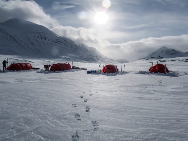 Røde telt satt opp i et snødekt fjelllandskap under en lys himmel med fotspor i forgrunnen.