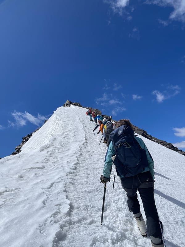 En rekke fjellklatrere med ryggsekker går langsomt oppover en bratt, smal snørygg som fører mot en steinete fjelltopp under en dyp blå himmel.