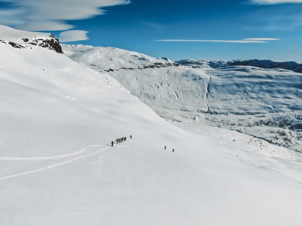 En gruppe skiløpere beveger seg i en rekke over et enormt, snødekt fjellandskap med hvite tinder og blå himmel i bakgrunnen.