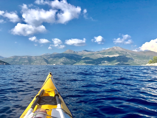 En førstepersonsvisning fra en gul kajakk på dypblått sjøvann, med utsikt mot en enorm fjellkjede under en klar, blå himmel med lettskyet vær.