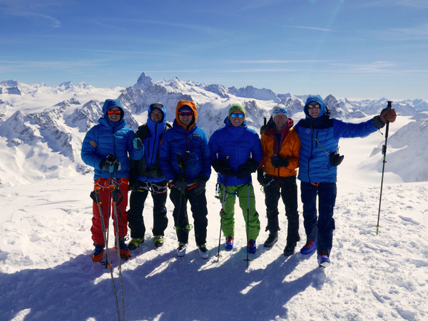 En gruppe på seks fjellklatrere i fargerike vinterklær og solbriller poserer sammen på en snødekt topp med et enormt panorama av alpine fjell i bakgrunnen.