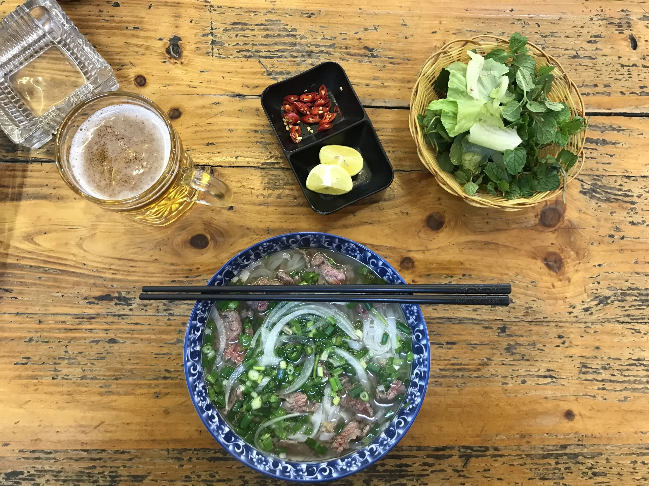 En bolle med pho med spiseptinner på et tresbord ved siden av et glass øl, chili, lime og friske urter.