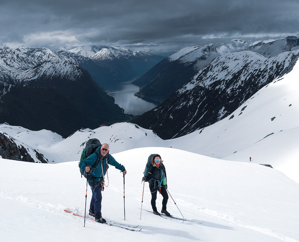 To skiløpere på en snødekt fjellrygg med en fjord og dramatiske snøfjell i bakgrunnen under en skyet himmel.
