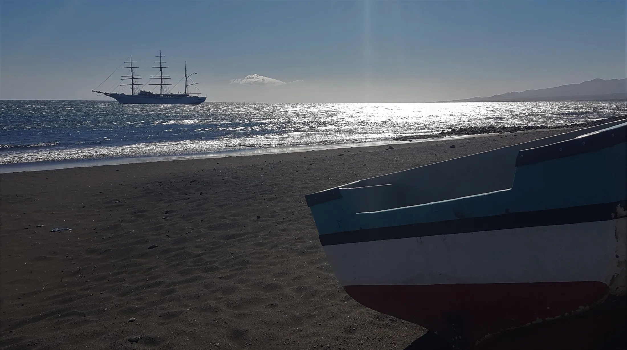 En trefiskerbåt hviler på en mørk sandstrand, med et stort fullrigget seilskip forankret i det glitrende, solfylte havet i bakgrunnen.