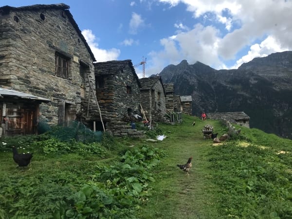 En rad med tradisjonelle steinhus ligger på en gresskledd skråning i en alpin landsby, med høner som går på stien og alpine fjelltopper som hever seg i bakgrunnen under en skyete himmel.