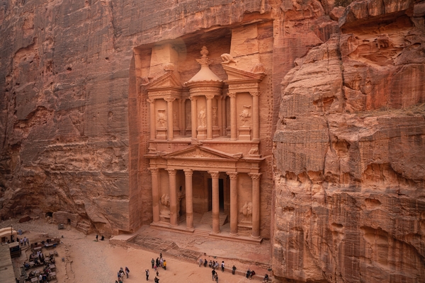 Den utskårne fasaden til Skattkammeret (Al-Khazneh) i Petra hugget direkte inn i den rødbrune sandsteinsveggen, med små turister synlige på bakken foran.