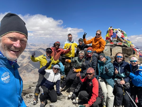 En smilende gruppe fjellklatrere poserer sammen på en steinete fjelltopp under en klar blå himmel, ved siden av en steinvarde dekket av fargerike bønneflagg.