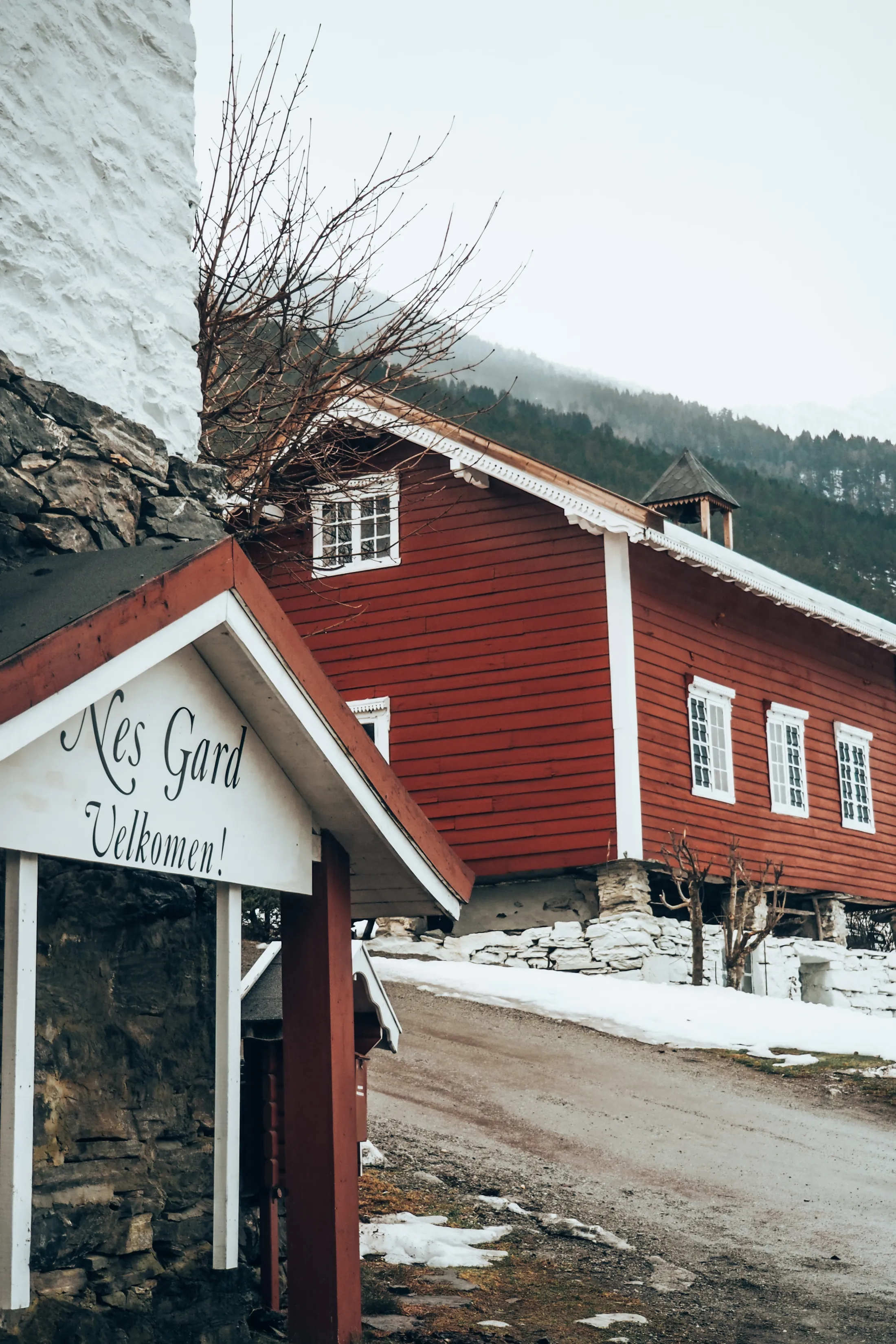 Et skilt med teksten «Nes Gard Velkomen!» står foran en tradisjonell rød låvebygning i et vinterlandskap med skogkledde fjell i bakgrunnen.