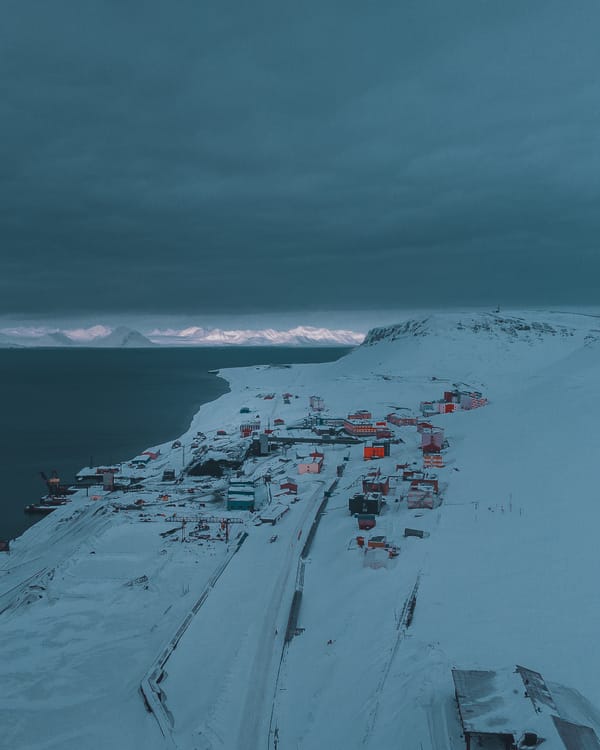 Et luftfoto av et snødekt kystsamfunn ved mørkt arktisk hav, med fjell opplyst av fjernt sollys under en tung, blågrå himmel.