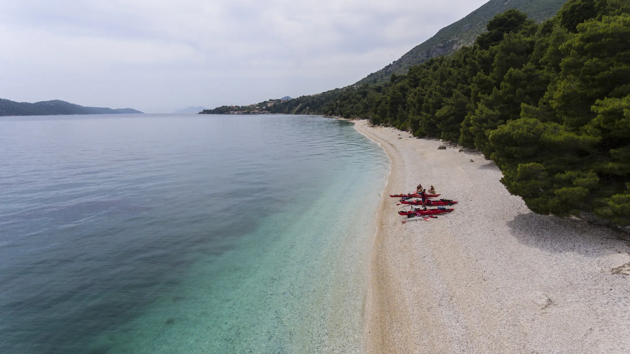 Et luftfoto i fugleperspektiv av flere røde kajakker og en liten gruppe mennesker som hviler på en lang rullesteinstrand ved siden av klart, lyst turkisblått vann som går over i et dypere blått hav, flankert av en frodig furuskog som strekker seg oppover en fjellside.