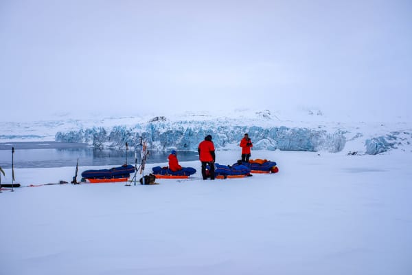Tre personer i røde vinterjakker hviler med de lastede pulkene sine på en snøfonn med utsikt over en stor, blå brevegg og åpent vann.