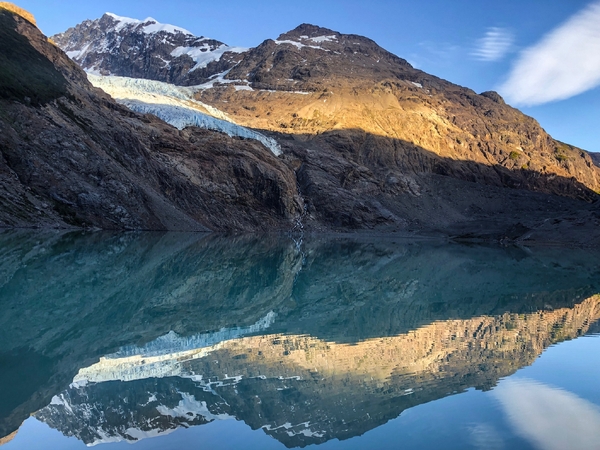 Speilbilde av et fjell med bre på i en klar turkis innsjø, under en blå himmel.