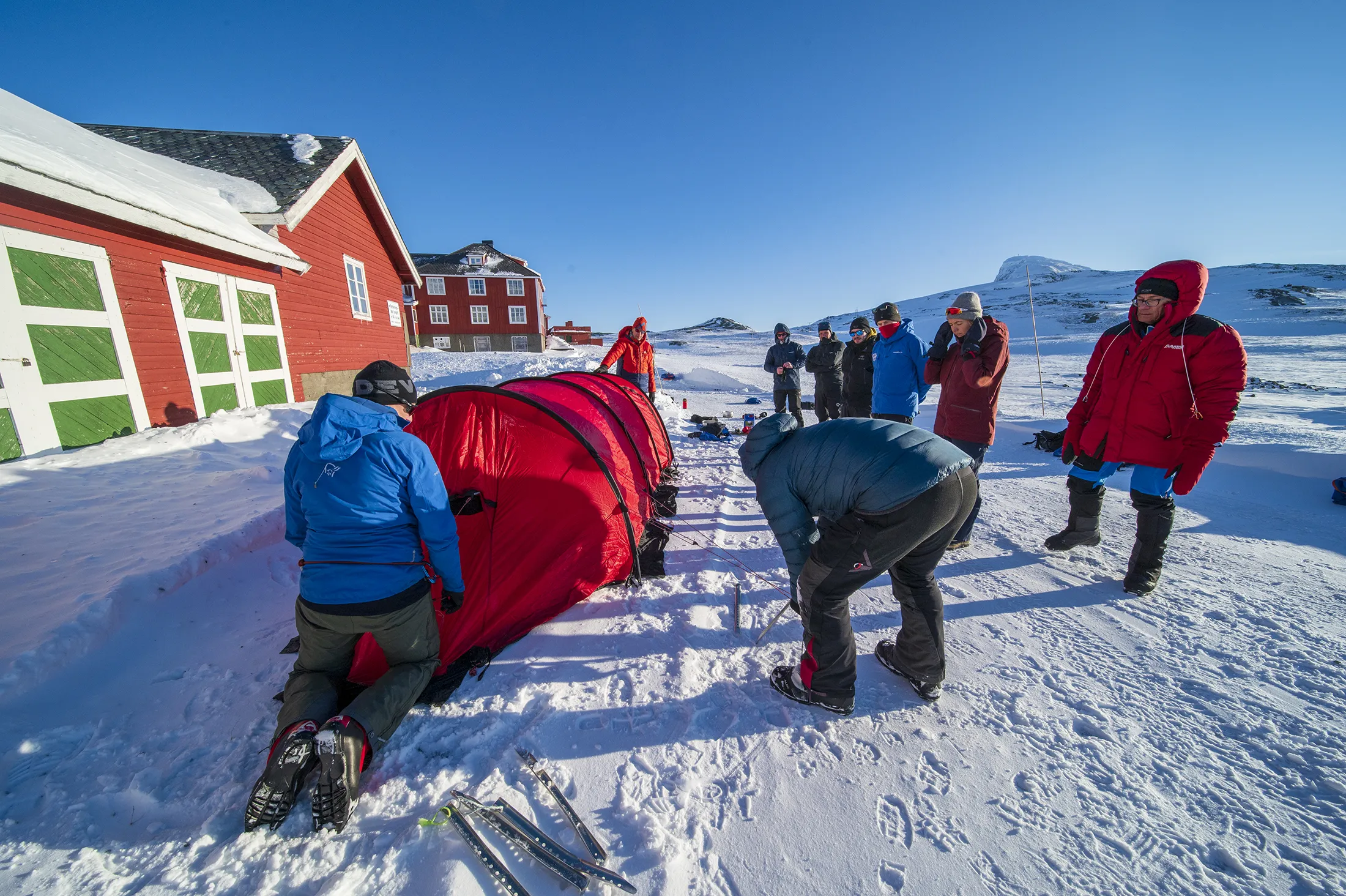 En gruppe mennesker som setter opp et rødt ekspedisjonstunneltelt i snøen utenfor tradisjonelle røde trebygninger på en solrik vinterdag.