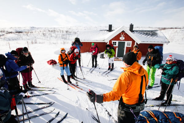 En gruppe fjellskiløpere samles utenfor en rød fjellhytte i snøen.