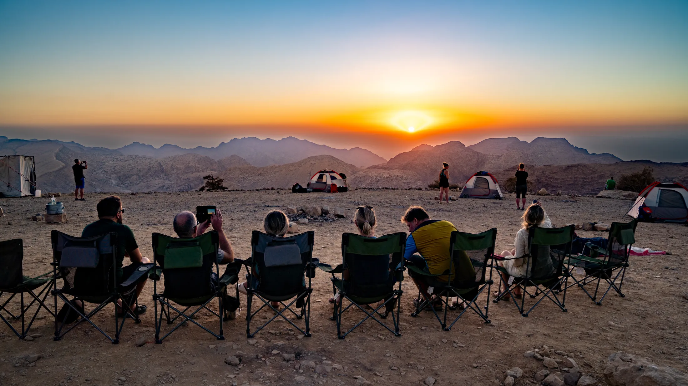 En gruppe turgåere sitter i campingstoler på rekke og ser ut over et vidstrakt fjellandskap under en fantastisk solnedgang.