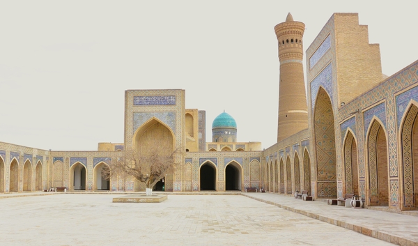 En stor, åpen gårdsplass er omkranset av historiske mursteinsbygninger med flislagte buer, et enslig, bart tre, en turkis kuppel og en høy minaret under en overskyet himmel.