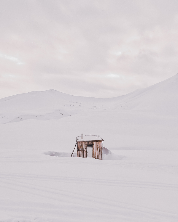 En liten, rustikk trehytte delvis begravd i dyp snø omgitt av et vaidt hvitt polarlandskap og fjell.