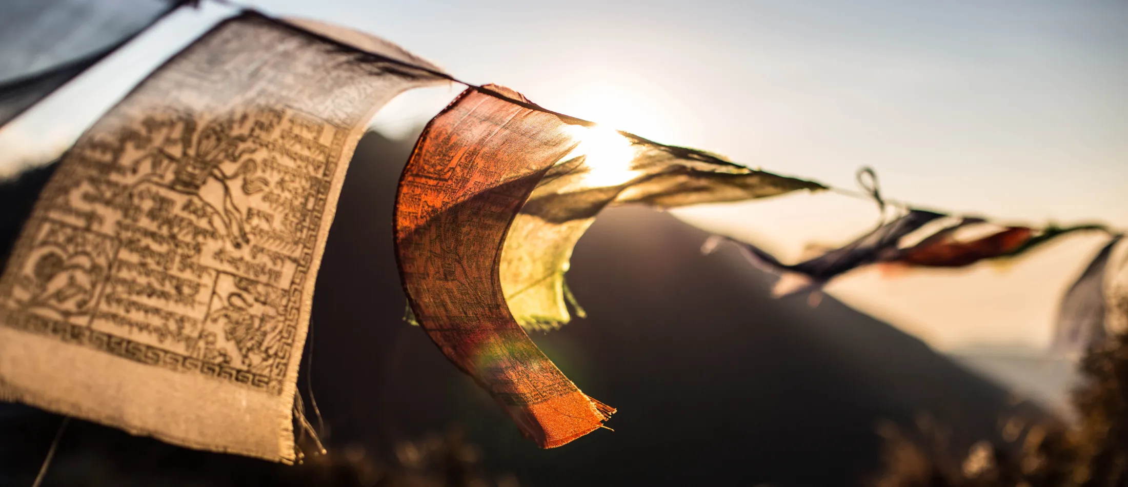 Et nærbilde fremhever fargerike, værbitte tibetanske bønneflagg som blafrer i vinden mens den sterke solen lyser rett bak dem og kaster et varmt lys mot en myk, uskarpt fjellbakgrunn.