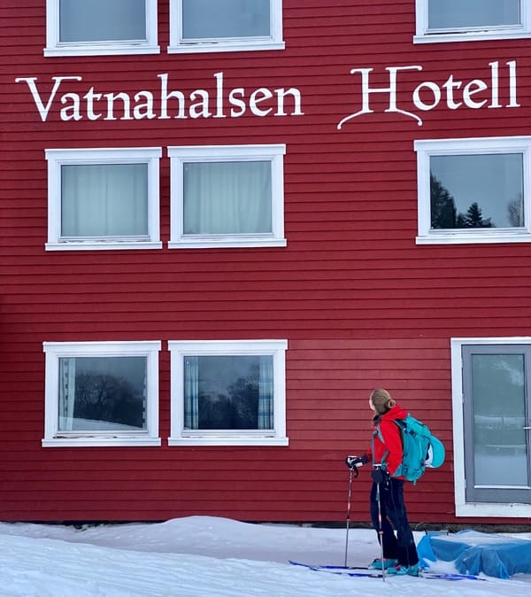 En person i skiutstyr står utenfor det rødmalte Vatnahalsen Hotell i snøen.
