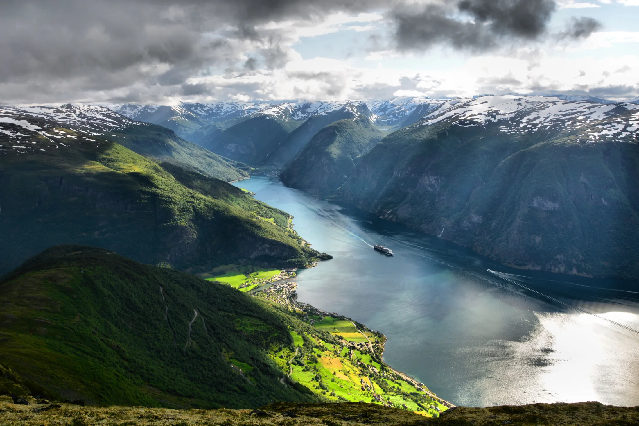 n fantastisk utsikt i fugleperspektiv over en dypblå fjord som svinger seg gjennom bratte, grønne fjell med snødekte topper, der et cruiseskip seiler forbi en liten bygd under en overskyet himmel.