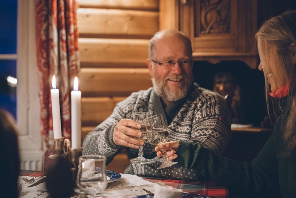 En eldre mann i mønstret ullgenser smiler mens han skåler med en annen person inne i en koselig tømmerhytte opplyst av levende lys.