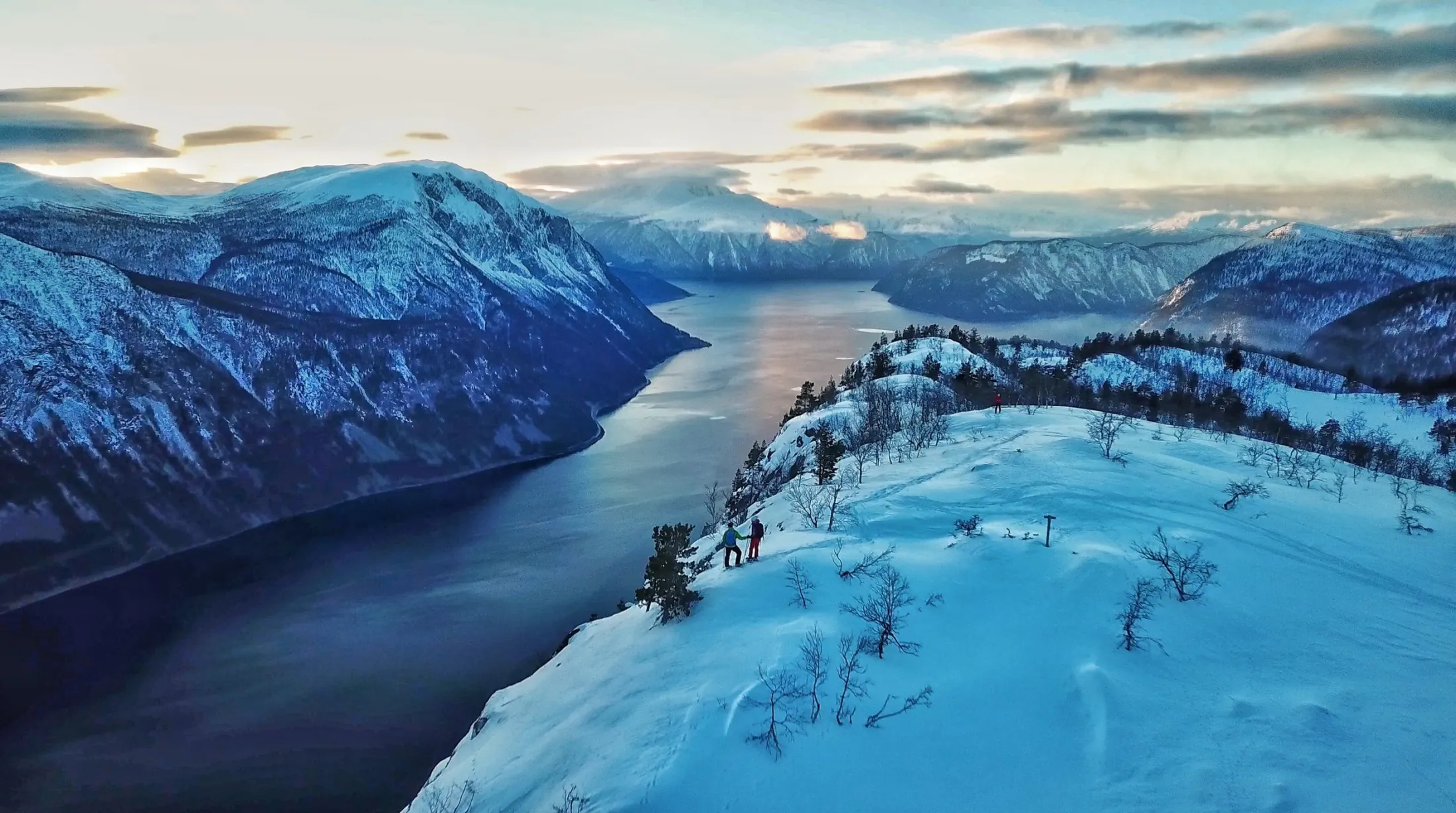Utsikt fra oven av to personer som står på en snødekt fjellrygg med utsikt over en dyp, blå fjord i skumringen.