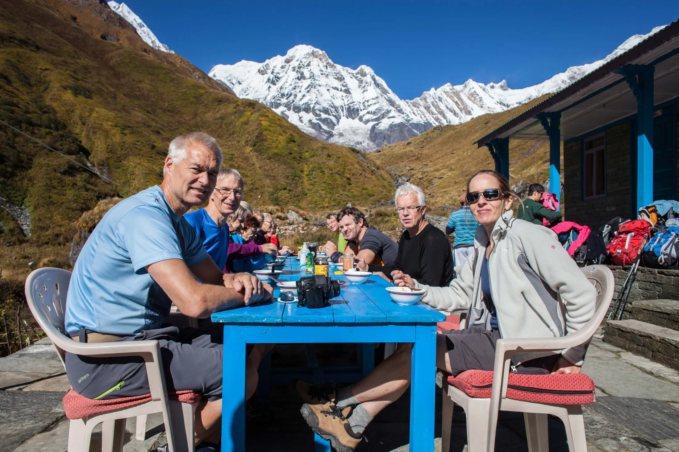 En gruppe turgåere sitter ved et langt, blått langbord og nyter et måltid utendørs, med en spektakulær bakgrunn av gylne åser og et massivt, snødekket fjellmassiv under en klar, blå himmel.