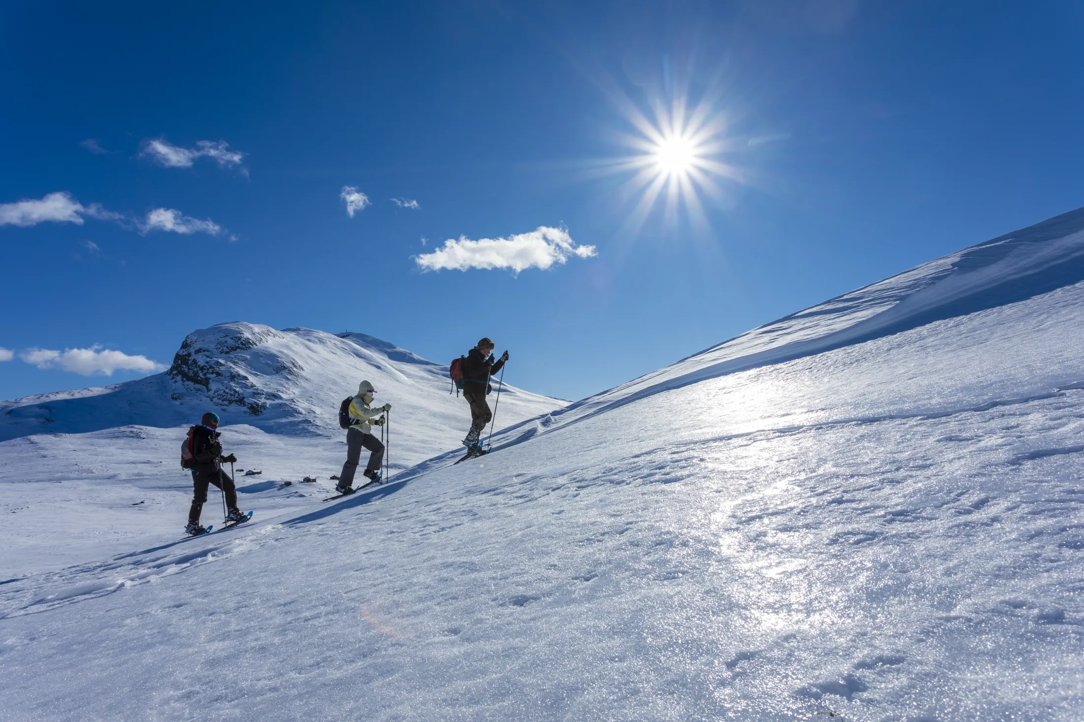 Tre personer med sekk og staver går i motbakke på truger over en snødekt fjellside under en lys, solfylt blå himmel.