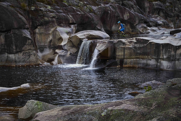 En terrengsyklist stående på toppen av en liten foss som renner ut i et mørkt, stille tjern omgitt av mosegrodde steiner i et kupert, fjellendt landskap.
