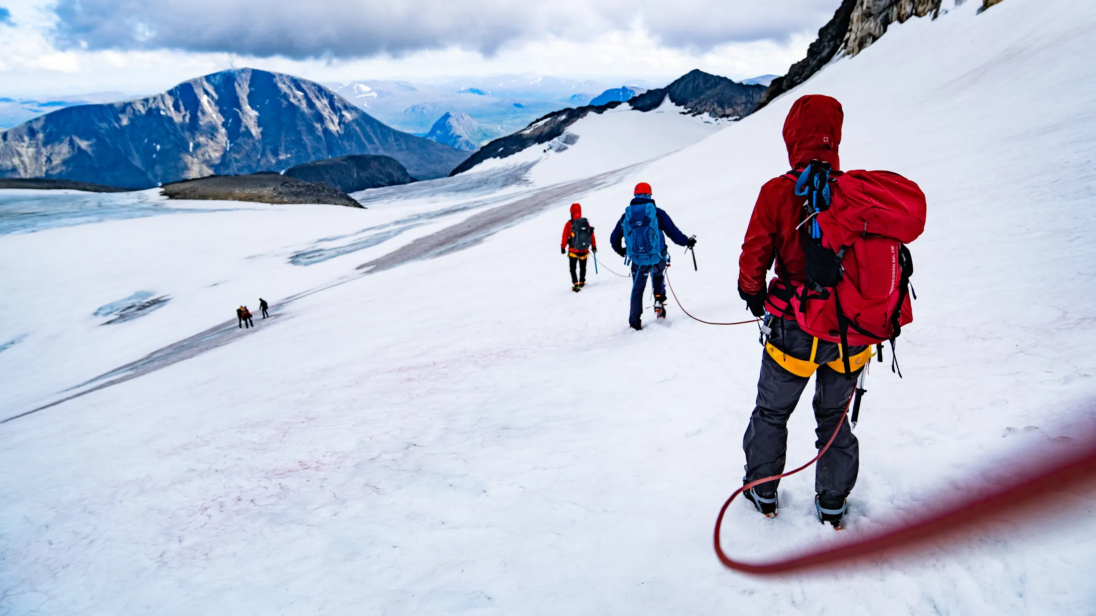 En gruppe tausaute fjellvandrere som går over et snødekt brelandskap under en overskyet himmel.