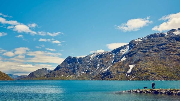 En person står på en steinete strand ved en blå innsjø med snødekte fjell i bakgrunnen under en delvis skyet himmel.