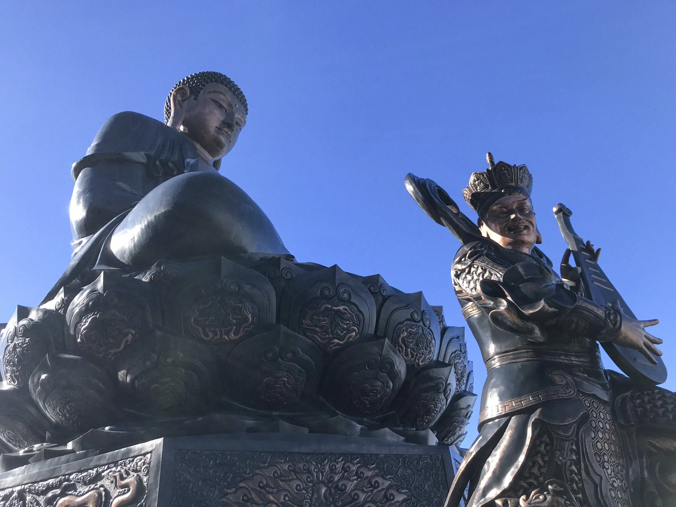 Et lavt vinkelbilde av to mørke bronsestatuer mot en klar blå himmel. Til venstre en sittende figur på en lotuslignende sokkel; til høyre en stående krigerfigur som holder våpen.