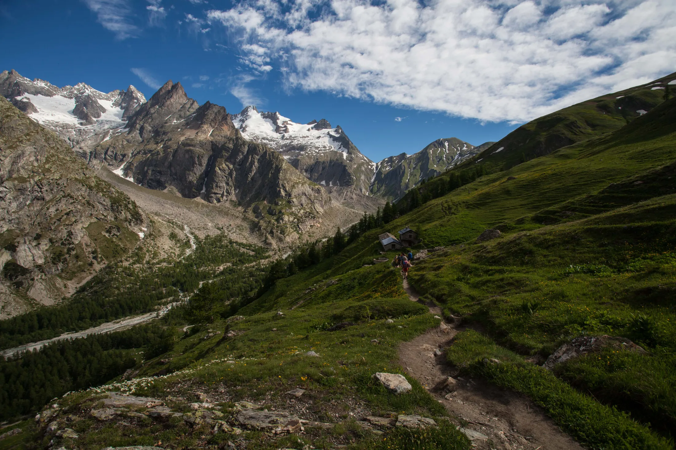 En smal sti svinger seg nedover en bratt, gresskledd fjellside mot små steinhytter, med majestetiske, snødekte fjelltopper i bakgrunnen under en blå himmel.