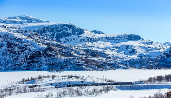 Et passasjertog kjører gjennom et snødekt fjellandskap forbi et frossent vann under en klar blå himmel.