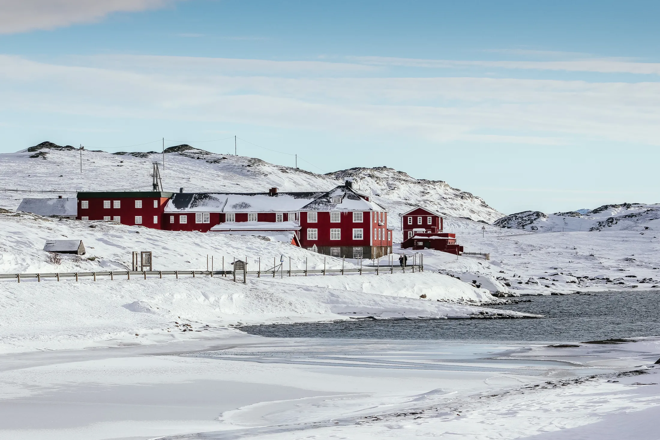 Tradisjonelle røde trebygninger ligger i et snødekt fjellandskap ved et delvis frossent vann under en klar blå himmel.