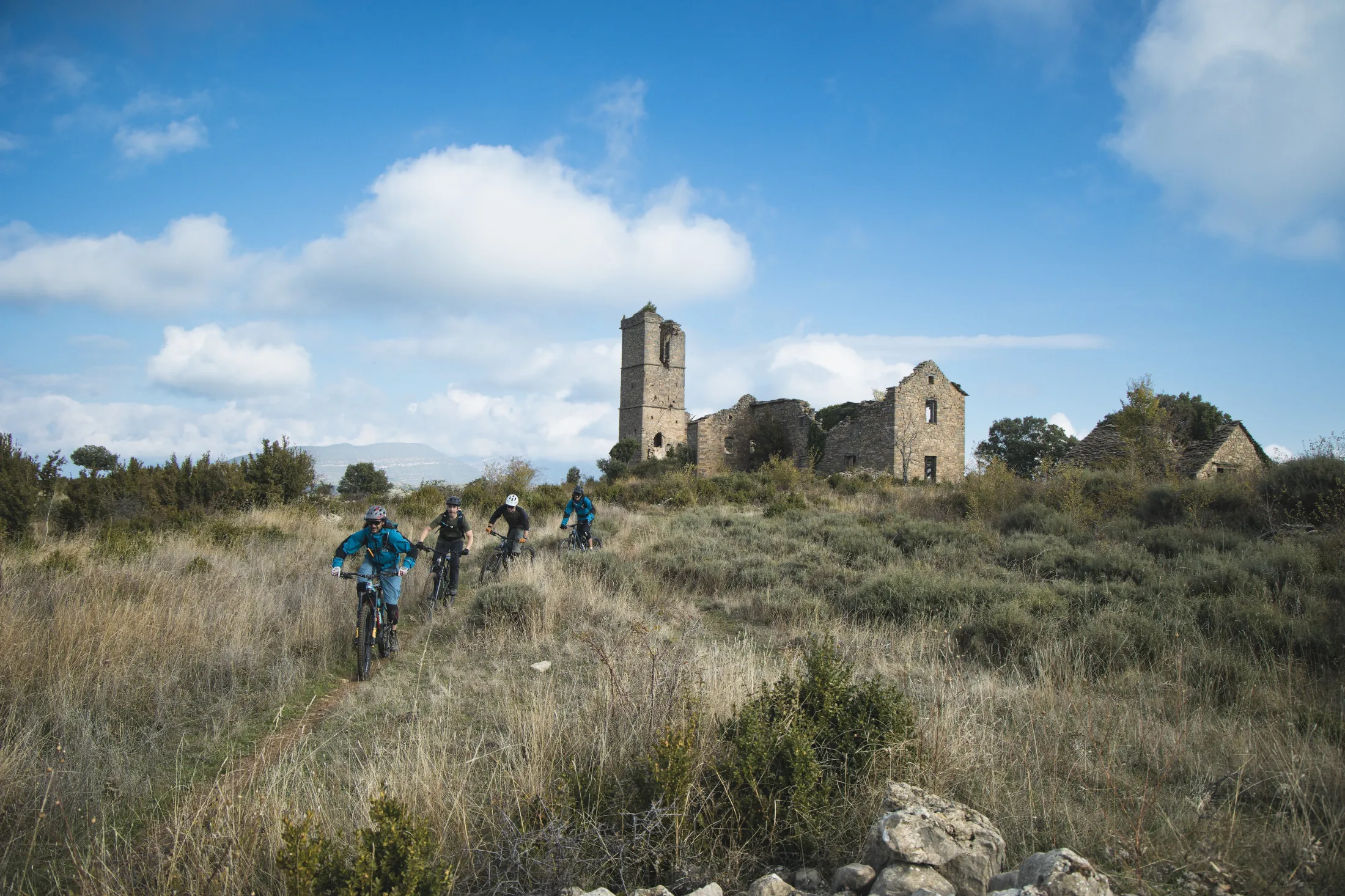 Terrengsyklister som sykler på en grussti mot en ruinert steinkirke under en blå himmel.