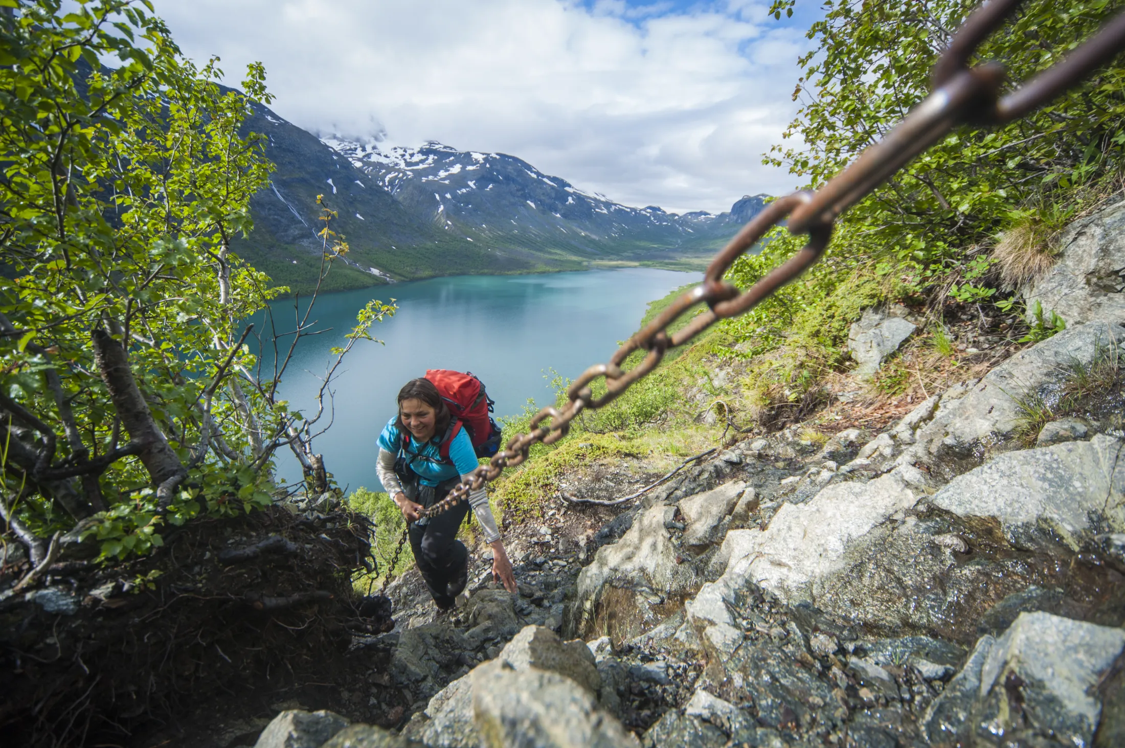 En turgåer med rød sekk går langs en bratt, steinete fjellsti sikret med en tung jernkjede, med utsikt over en turkis fjord og snødekte fjell.